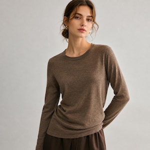 May include: A long-sleeved crew neck sweater in a solid medium brown color. The garment features a fine-knit texture, a classic round neckline, and fitted sleeves, providing a simple and clean silhouette for everyday wear in a neutral tone.