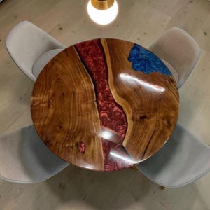 May include: A round wooden dining table featuring a central river of swirling red resin and a smaller accent of blue resin. The table is surrounded by four light grey upholstered chairs, viewed from directly above under a single pendant light.