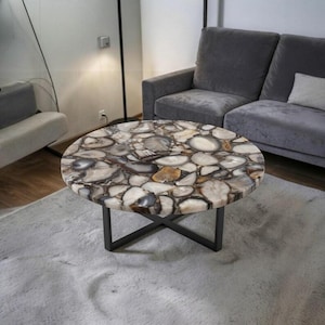 Puede incluir: Una mesa de centro redonda con un tablero compuesto por numerosas rodajas de ágata pulidas y multicolores en tonos blanco, gris, marrón y beige. La mesa está sostenida por una base de metal negro con un diseño de patas en cruz.