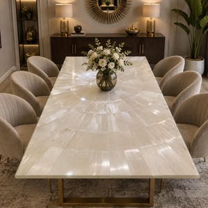 May include: A rectangular dining table with a white, fan-patterned stone surface and a gold metal base, surrounded by six beige upholstered chairs with vertical channel tufting and gold legs, featuring a glass vase with white flowers.