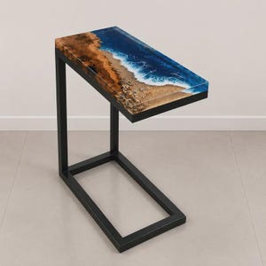 May include: A C-shaped side table with a black metal frame and a rectangular tabletop. The tabletop features a natural wood edge combined with a blue resin inlay that mimics a coastal shoreline with sandy beaches and white ocean waves.