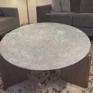 Peut inclure: Une table basse ronde dotée d'un plateau en pierre blanche translucide aux multiples facettes, présentant un aspect en mosaïque. La table repose sur deux pieds épais et incurvés en bois brun foncé qui forment une base solide pour la surface.