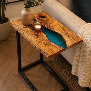 Puede incluir: Una mesa auxiliar en forma de C con un tablero de madera natural de borde irregular y una incrustación central de resina turquesa vibrante. La mesa se apoya sobre una base sólida de marco de metal negro con un diseño rectangular.