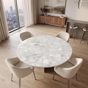 Puede incluir: Una mesa de comedor redonda con un tablero de aspecto de piedra blanca y gris con un patrón fragmentado estilo mosaico. La mesa está rodeada por cuatro sillones tapizados en color beige claro con respaldos curvos y patas metálicas finas.