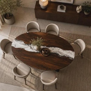 May include: An oval dining table featuring a dark brown wood surface with a central white and grey marble inlay, surrounded by six beige upholstered chairs. A small round tray with two dark bowls sits in the centre of the table.