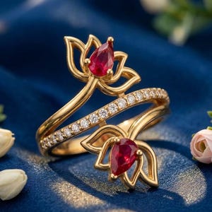 Könnte beinhalten: Ein goldener Wickelring mit zwei offenen Lotusblütenmotiven, die jeweils mit einem birnenförmigen roten Edelstein besetzt sind. Ein schmales Band, das mit einer Reihe kleiner, klarer, rund geschliffener weißer Steine besetzt ist, verbindet die Blüten.