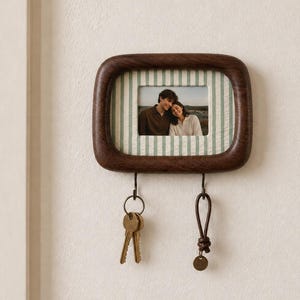 May include: A dark brown wooden wall-mounted photo frame with rounded corners and a light green and white striped mat. The frame holds a photograph of two people and features two metal hooks at the bottom, each holding a set of metal keys.