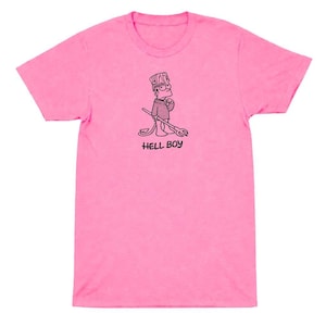 May include: A bright pink short-sleeved t-shirt featuring a black line art graphic of a cartoon character with spiky hair, a tail, and a trident. The text HELL BOY is printed in black capital letters directly underneath the illustrated character.