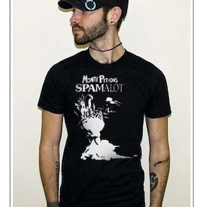 Può includere: Una t-shirt nera a maniche corte con la scritta bianca Monty Python's SPAMALOT e una grafica bianca di un elmo con pennacchio. La maglietta è abbinata a un berretto nero con un piccolo emblema circolare blu e bianco sul lato.