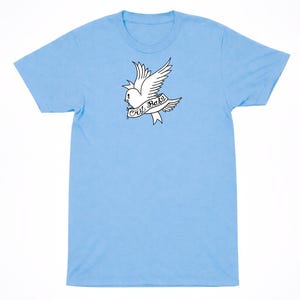 May include: A light blue short-sleeve t-shirt featuring a black and white graphic of a dove in flight. The bird has a teardrop under its eye and carries a banner across its chest with the text Cry Baby written in a cursive script font.
