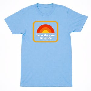 May include: A light blue short-sleeve t-shirt featuring a centered graphic of a yellow, orange, and red semi-circle rainbow inside a rounded orange square. The text "hawthorne heights" is printed in white, lowercase, serif font below the rainbow.