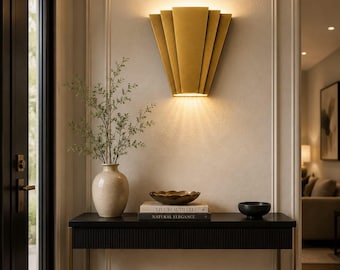 May include: A gold-toned wall sconce with a fan-shaped design mounted above a black console table. The table holds a beige ceramic vase with greenery, a shallow gold bowl, and two books titled Live Beautifully and Natural Elegance.