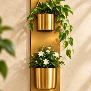 Pode incluir: Uma placa de parede retangular vertical de metal dourado com dois vasos cilíndricos dourados. O vaso superior contém uma planta pothos variegada verde e amarela, enquanto o inferior abriga um pequeno arbusto com flores brancas.