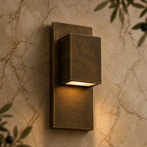 May include: A rectangular wall sconce with a dark bronze finish, featuring a vertical backplate and a protruding box-shaped light housing that casts a warm, downward-facing glow against the wall surface.