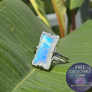 May include: A silver ring featuring a rectangular moonstone with a blue iridescent sheen, held in a four-prong setting. A circular badge in the corner displays the text: FREE COLLECTABLE STONE WITH ANY PURCHASE! with a small purple crystal icon.