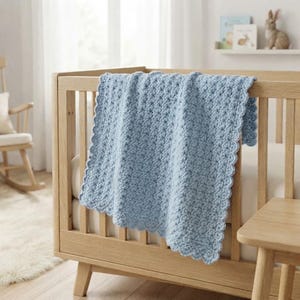 May include: A light blue crocheted baby blanket with a textured stitch pattern and a scalloped edge draped over the side of a light-colored wooden crib. The blanket features a consistent yarn pattern throughout and a decorative border.