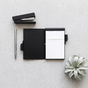 Puede incluir: Una grapadora negra, un bolígrafo de metal plateado y un cuaderno negro abierto con páginas blancas. El cuaderno tiene una cubierta negra y una trabilla para el bolígrafo. Los artículos están dispuestos sobre una superficie gris claro moteada.