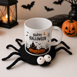 May include: A white ceramic mug featuring a black graphic of a haunted house, pumpkins, bats, a spider web, and the text Happy Halloween, resting on a black crocheted spider-shaped coaster with two white crocheted eyes attached to the front.