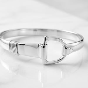 Op de afbeelding: Een gepolijste zilverkleurige armband met een centraal, op de paardensport geïnspireerd stijgbeugelmotief. De band heeft aan één kant een glad, afgerond profiel en aan de andere kant een plat, breed gedeelte, geaccentueerd met fijne draaddetails.