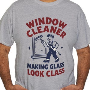 Könnte beinhalten: Ein graumeliertes Kurzarm-T-Shirt mit einer Grafik eines Fensterreinigers, der einen Abzieher hält. Der Text lautet "WINDOW CLEANER" in Dunkelrot und "MAKING GLASS LOOK CLASS" in Dunkelblau, ergänzt durch eine kleine Fenstergrafik.