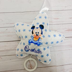 Puede incluir: Un cojín de peluche con forma de estrella en tela blanca con un patrón de estrellas azules. El centro muestra un personaje de ratón de dibujos animados y el nombre "Cristian" en letra azul. Tiene una cinta blanca arriba y un anillo de plástico.