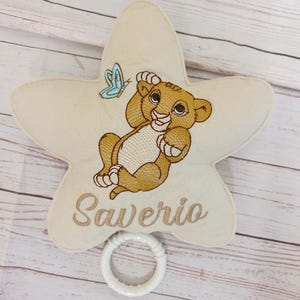 Pode incluir: Um ornamento de tecido cor de creme em forma de estrela com um filhote de leão castanho bordado e uma borboleta azul clara. O nome Saverio está bordado em letra cursiva castanha abaixo do leão. Um anel de plástico branco está preso embaixo.