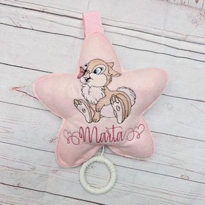 Può includere: Un peluche rosa chiaro a forma di stella con un coniglio ricamato marrone e bianco che ha una piccola farfalla rosa sul naso. Il nome Marta è ricamato in corsivo rosa scuro sotto il coniglio, con un anello in plastica bianca attaccato.