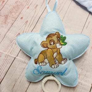 Può includere: Un peluche azzurro a forma di stella con un leoncino marrone ricamato e una farfalla verde sul naso. Il nome Samuel è ricamato in blu sotto il leone. Un anello di plastica bianca è fissato sul fondo e un nastro azzurro si trova sulla parte superiore.