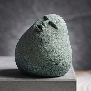 May include: A smooth, rounded, sage green stone sculpture featuring a stylized human face with closed eyes and a small, pursed mouth. The object has a textured, matte surface and a teardrop-like shape, resting on a flat, rectangular base.