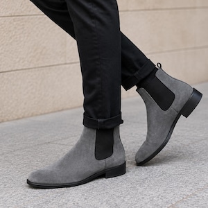 Puede incluir: Un par de botines Chelsea de ante gris con paneles laterales elásticos negros, una trabilla en el talón y un tacón bajo negro. Los botines se llevan con pantalones negros con dobladillo, mostrando un diseño elegante con punta redondeada.