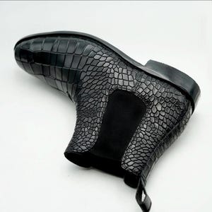 Könnte beinhalten: Ein schwarzer Stiefeletten-Schuh mit einer strukturierten Krokodilprägung auf dem Obermaterial. Der Schuh verfügt über einen einfarbigen schwarzen elastischen Seiteneinsatz, eine abgerundete Zehenpartie, einen niedrigen Absatz und eine Zuglasche.