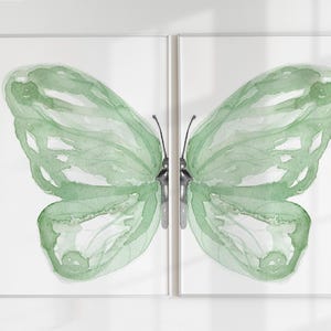 May include: A diptych wall art set featuring a watercolour painting of a butterfly split across two separate framed panels. The butterfly wings are rendered in varying shades of sage green with translucent, fluid brushstrokes and thin antennae.