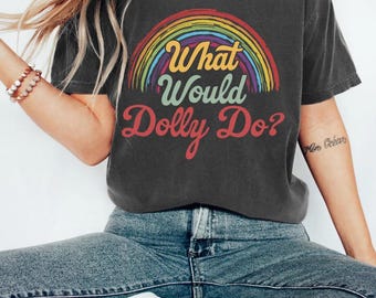 May include: A charcoal grey short-sleeve t-shirt featuring a colourful rainbow graphic arched over the text "What Would Dolly Do?" in a mix of yellow, light green, and red script and serif fonts. The shirt has a crew neckline and a relaxed fit.