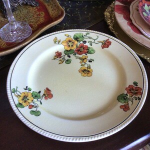 May include: A white ceramic dinner plate with a yellow, orange, and green floral design. The plate has a scalloped edge and a raised rim.