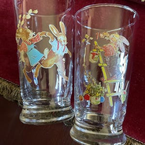 May include: Two clear glass tumblers with colorful Easter bunny illustrations. One tumbler features bunnies dancing, the other shows a bunny painting. The glasses have a flared base and straight sides, perfect for serving beverages.