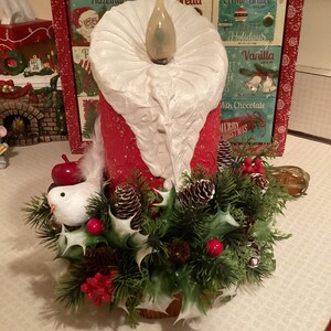 May include: A red and white candle with a white wax drip, surrounded by a green garland with pine cones, holly, and red berries. A small white bird with a red apple sits on the garland. The candle has a light bulb inside.