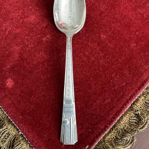 May include: A silver souvenir spoon with a detailed image of a tall building on the spoon's bowl. The handle is engraved with "NEW YORK" and "1932". The spoon is displayed on a red velvet surface.