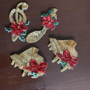 May include: Four gold sequined musical-themed Christmas ornaments. The ornaments include a treble clef, a musical note, and two bell shapes. Each ornament features a red sequined bow and accents of green and white.