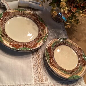 May include: A set of vintage plates with a cream-colored center and a decorative border featuring a landscape design in shades of brown, green, and gold. The plates are arranged on a white lace-trimmed tablecloth.