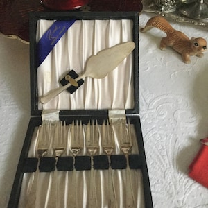 May include: A vintage silverware set in a black box with a blue ribbon. The set includes a cake server and six forks, all with silver-colored metal. The box is lined with white satin fabric.
