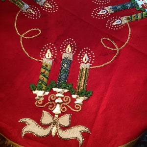 May include: A red Christmas tree skirt with gold fringe and embroidered candles. The candles are green, blue, and gold with white flames and are embellished with sequins. A gold bow and candle holder are also featured.