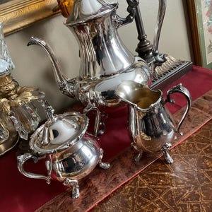 May include: A silver tea set with a teapot, creamer, and sugar bowl. The set has an ornate design with a floral motif. The pieces are all polished and shiny.