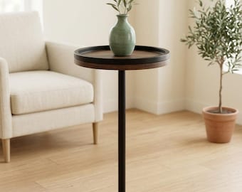 May include: A round side table featuring a dark wood top with a raised edge, a slender black metal central post, and a matching dark wood circular base. A small, light green ceramic vase containing green sprigs sits on the table.