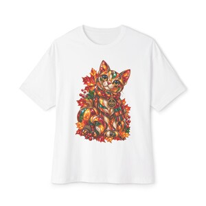 Może przedstawiać: Biały t-shirt z krótkim rękawem z kolorową grafiką kota w szaliku, otoczonego jesiennymi liśćmi, jagodami, dynią i żołędziami. Okrągły wisiorek zawieszony na szaliku zawiera napis 999 CLUB umieszczony w centralnej części grafiki.