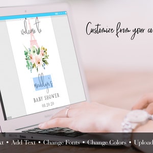 May include: A laptop displays a baby shower invitation design. The invitation features a watercolor Eiffel Tower, floral accents, and the text "Welcome to Madelyn's Baby Shower 08.20.20." The screen also shows editing options: Edit Text, Add Text, Change Fonts, Change Colors, and Upload Images.