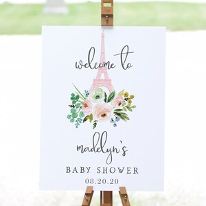 May include: A white sign on a wooden easel with the text "welcome to madelyn's BABY SHOWER 08.20.20". The sign features a pink Eiffel Tower illustration and a floral bouquet with pink, green, and yellow flowers.