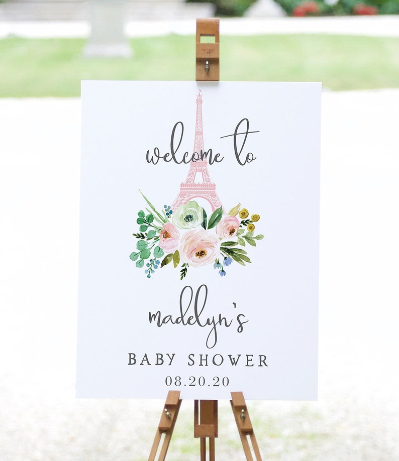 May include: A white sign on a wooden easel with the text "welcome to madelyn's BABY SHOWER 08.20.20". The sign features a pink Eiffel Tower illustration and a floral bouquet with pink, green, and yellow flowers.