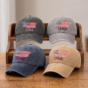 Könnte beinhalten: Vier Baseballkappen in Anthrazit, Hellgrau, Marineblau und Beige, jeweils mit einer gestickten amerikanischen Flagge und dem Schriftzug USA auf der Vorderseite. Die Kappen haben ein verwaschenes Design mit einem gebogenen Schirm.