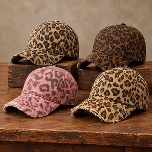 May include: Four baseball caps featuring a leopard print pattern. Three caps have a tan and brown color scheme, while one cap has a pink and brown color scheme. Each cap has a distressed, frayed edge along the brim and a six-panel construction.
