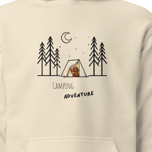 May include: A cream-colored hooded sweatshirt featuring a black line art graphic of a tent, pine trees, a crescent moon, and a small brown dog inside the tent. Below the graphic, the text reads Camping ADVENTURE in a simple black font.